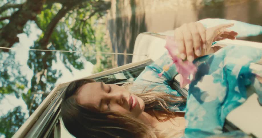 A woman in a blue floral robe leans out of a car window holding a flower appearing calm and serene