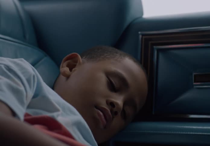 A young black child lies in the back seat of a moving car at midday sleeping throughout the journey
