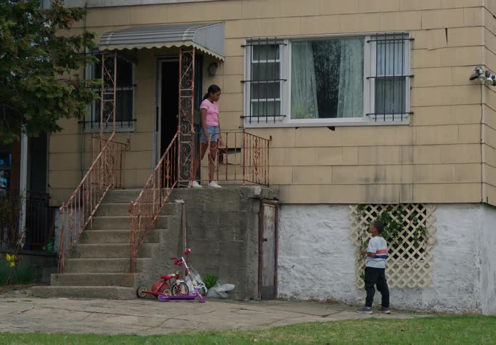 A young male kid plays with a string outside a building as a teenage girl walks out the door talks to him briefly then walks back inside