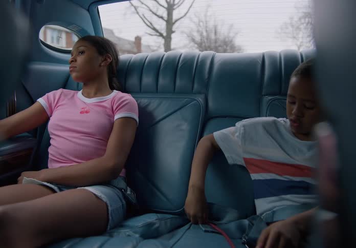 Two kids sit in a vintage blue car one staring out the window the other playing with a red strap