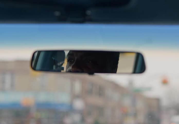 A mans focused eyes are visible in the rearview mirror as he drives through a city street