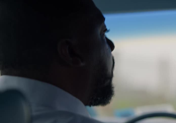 A person with short dark hair and a beard wearing a lightcolored shirt is seen from behind the passenger seat while driving a vehicle