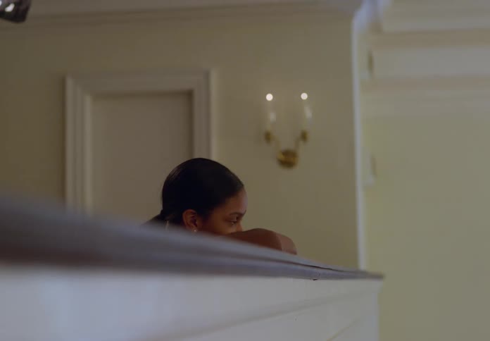 A child with short dark hair rests their arms on a white railing looking to the right in an indoor setting with lightcolored walls and a sconce