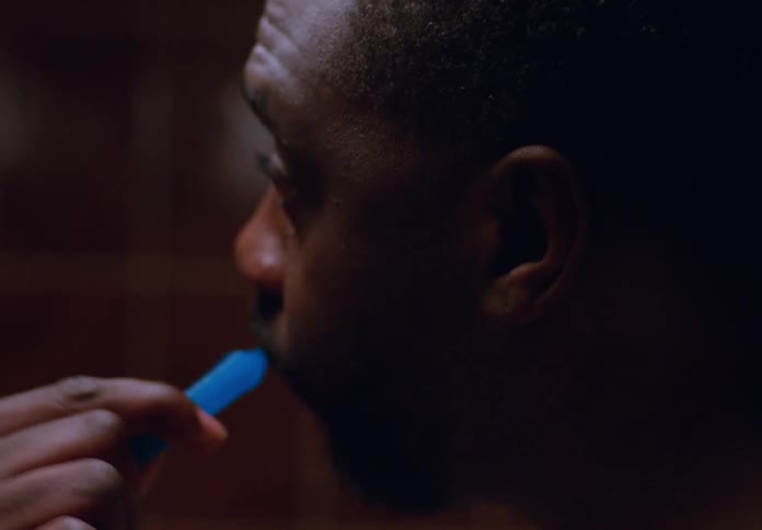 A person with short dark hair and a beard is seen in profile brushing their teeth with a blue toothbrush in a dimly lit setting