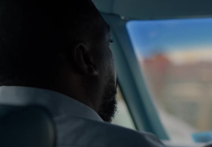 A black man wearing a colorful shirt sits behind the wheel driving a car at midday