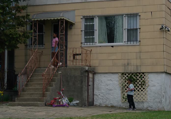 In a residential neighborhood at midday a child plays outside a home while his teenage sister walks out talks to him and then walks back inside as the child ignores her and continues his activity
