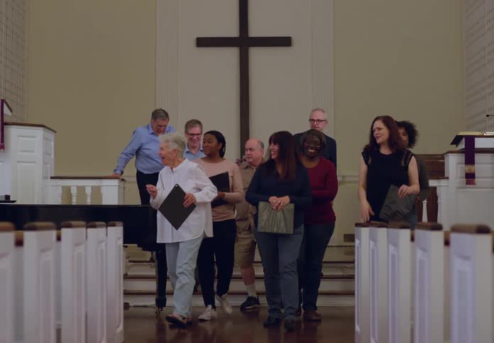 A choir sings in a church while a conductor leads them in front of a large cross