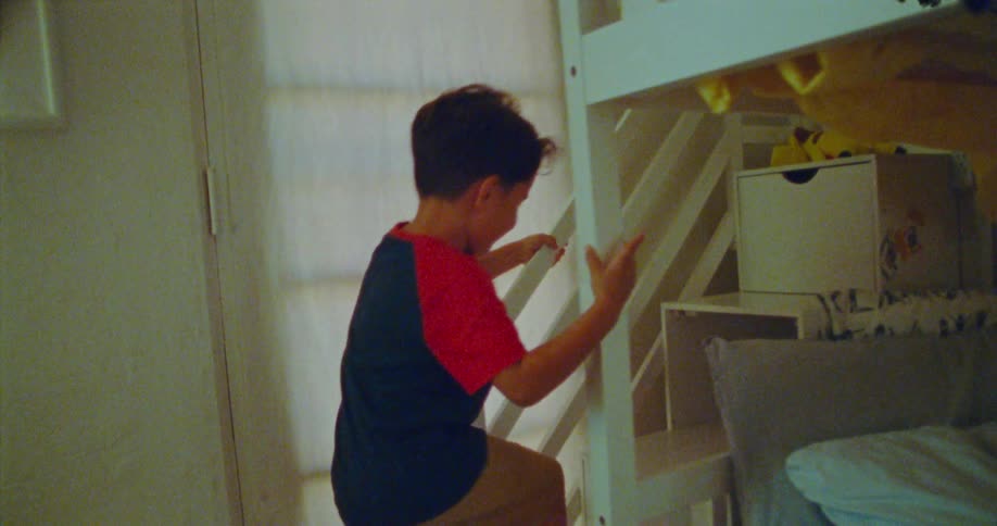 A little boy walks to his bunkbed and climbs on a ladder to the top bunk