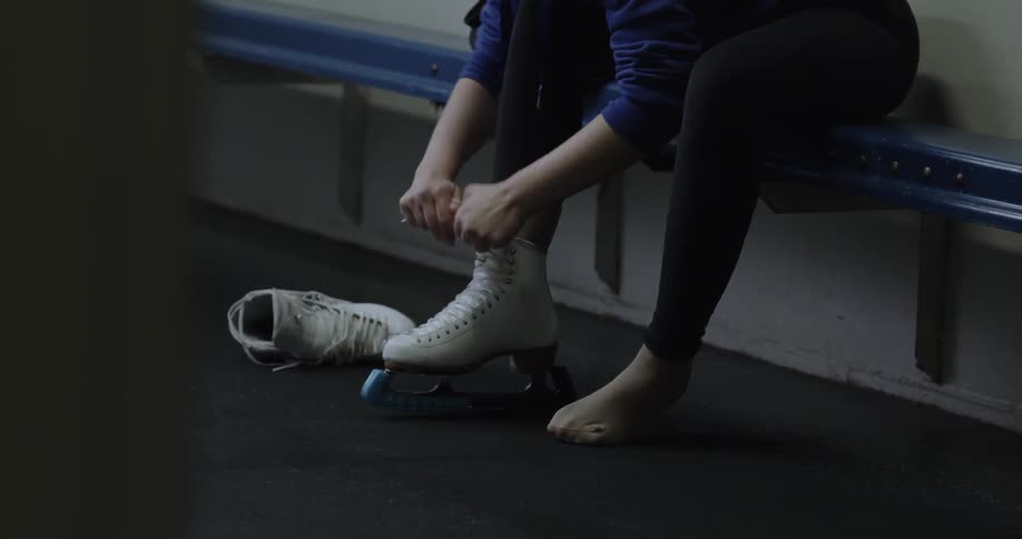 Blonde woman with a ponytail lacing up one of her ice skates while sitting on a bench