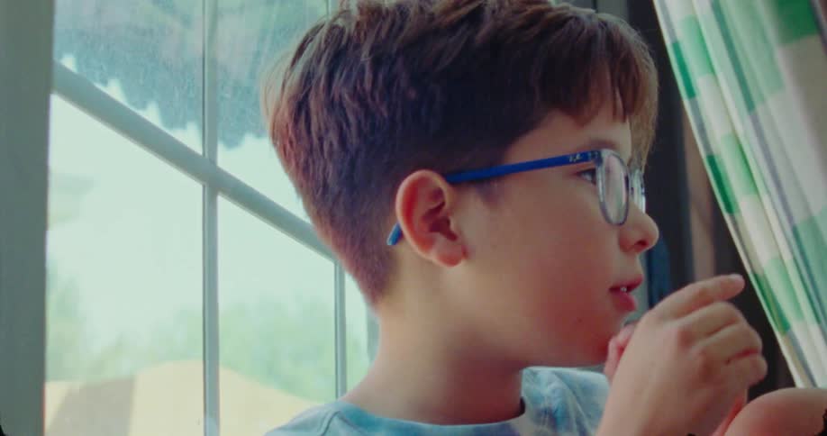 A young boy wearing glasses hides behind a curtain next to the window at midday covering his mouth to stay quiet