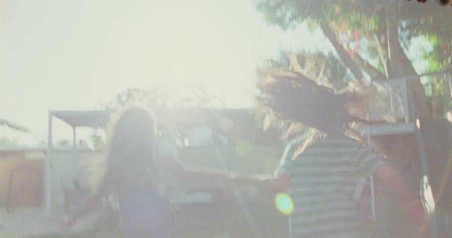 Two girls with long hair are holding hands and running through a sundrenched yard with bright pink flowers