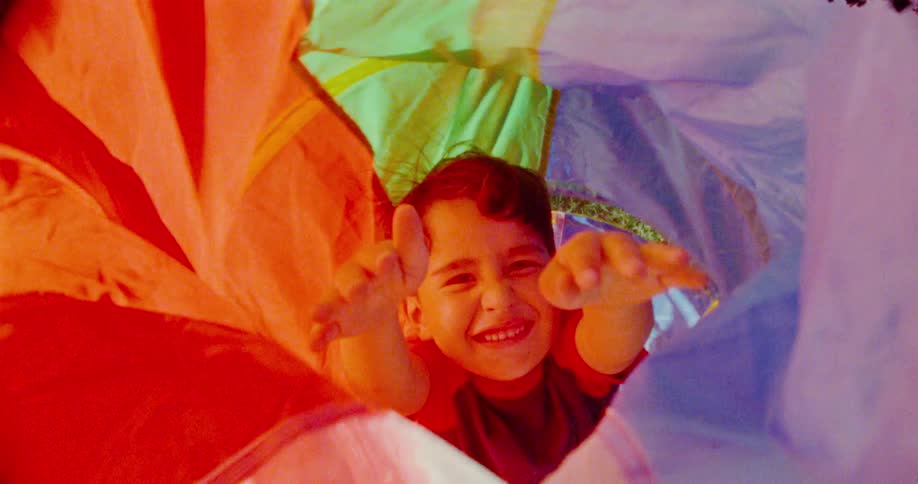 A smiling child with dark hair extends his arms forward from inside a colorful play tunnel