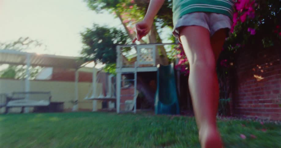 A child with bare feet and shorts is seen running through a grassy backyard towards a vibrant pink flowering bush