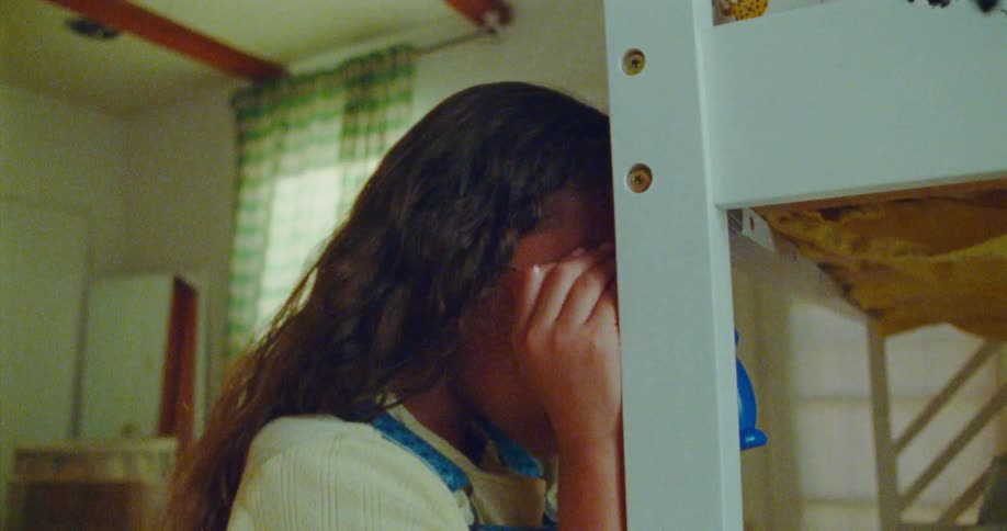 A young girl wearing glasses hides her face on the side of a bunk bed playfully calling out for a game of hide and seek
