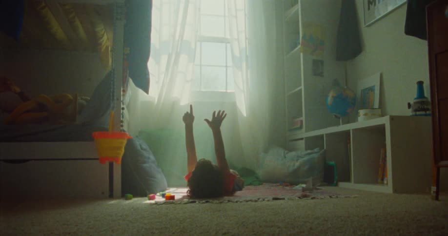 A child lies on the floor inside a room beside a window at midday with both hands raised up and surrounded by toys