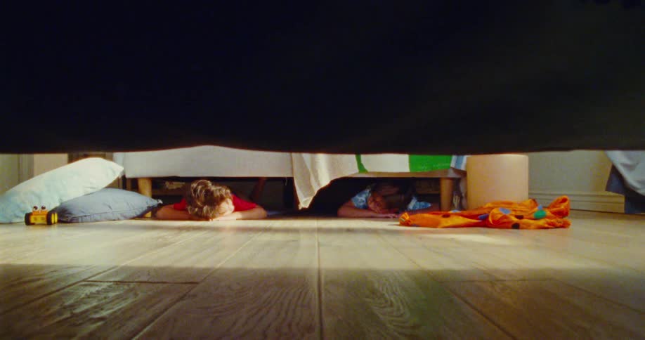 Two children are lying on a wooden floor hiding under a bed with scattered toys and clothes playing hideandseek