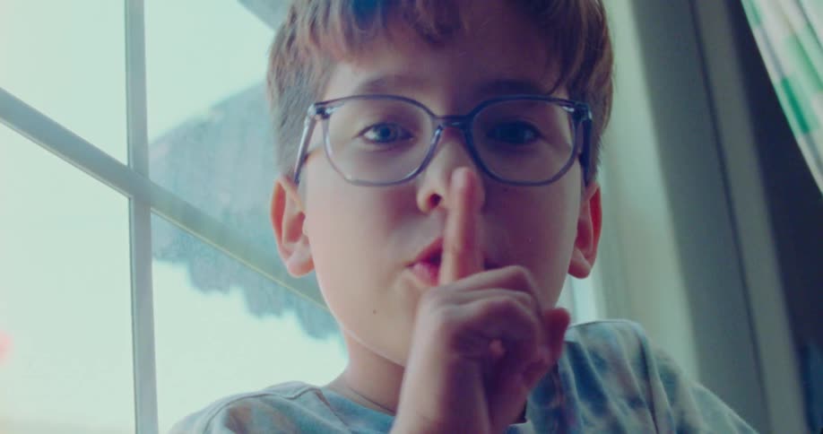 Boy hides by window whispering with finger on lips during hide and seek