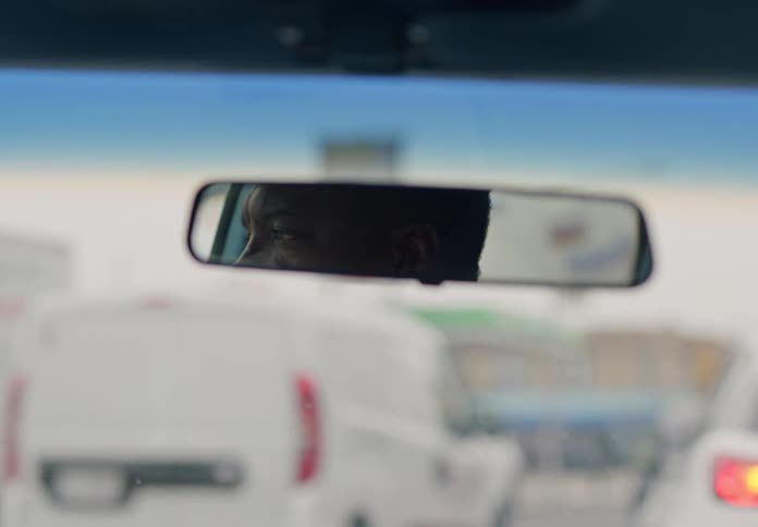 A black man is seen in the rearview mirror inside a car with a series of cars passing by on the road visible through the windshield at midday