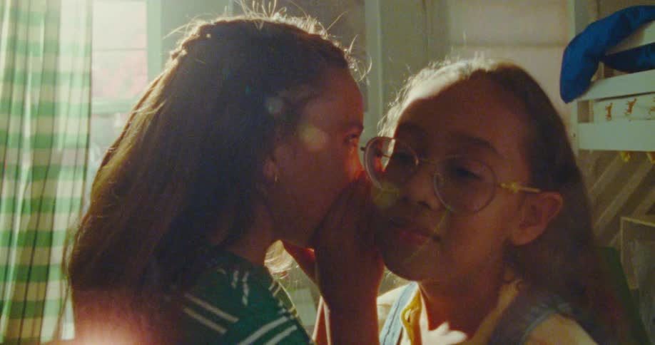A female child whispers into the ear of another girl wearing glasses and both leave the room afterward