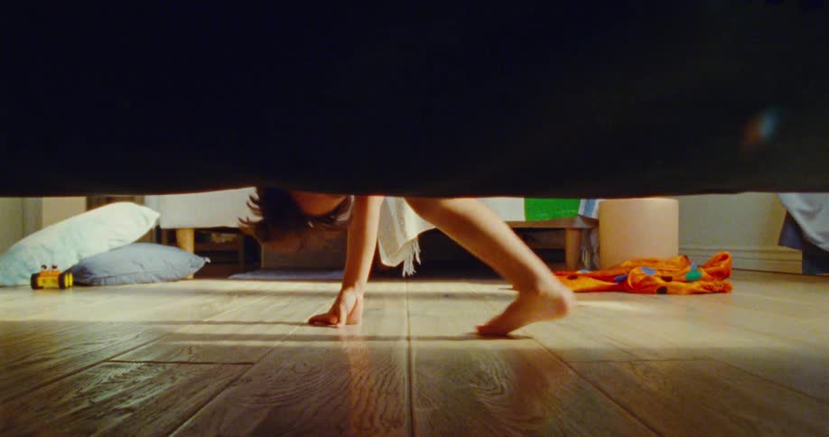 A barefoot child playfully climbs down from a bed onto a wooden floor in a sunlit room