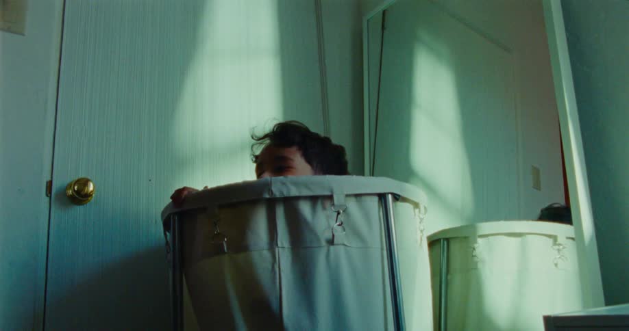 A child with dark curly hair peeks over the edge of a round lightcolored laundry hamper in a room with white doors and a mirror