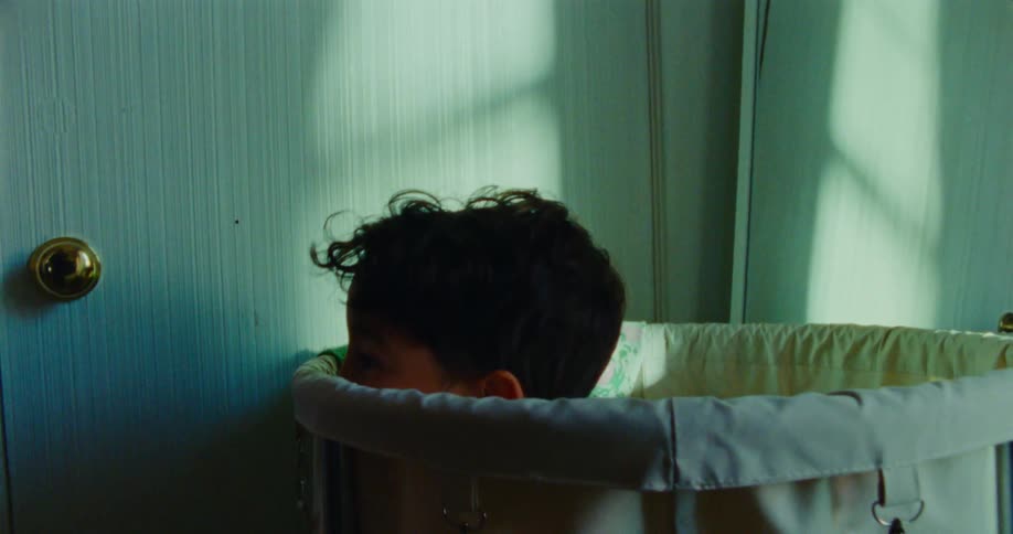 A child with dark curly hair is hiding inside a portable crib in a room with sunlight casting shadows on a white door