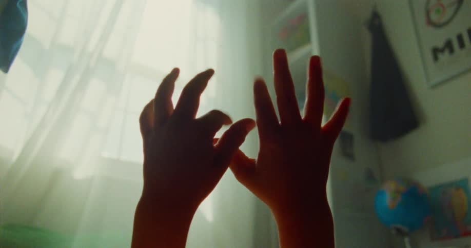 Child raises their hands and wiggles their fingers while sun shines into the room through a curtained window