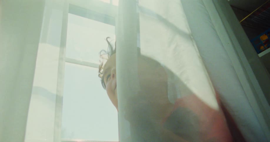 A kid hides behind a curtain by a window at midday during a game of hide and seek
