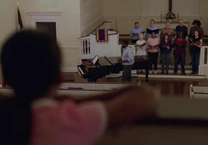 A group of people stand on a stage singing as a choir with a black man in front leading them he later walks toward a grand piano while a little girl in the audience watches attentively