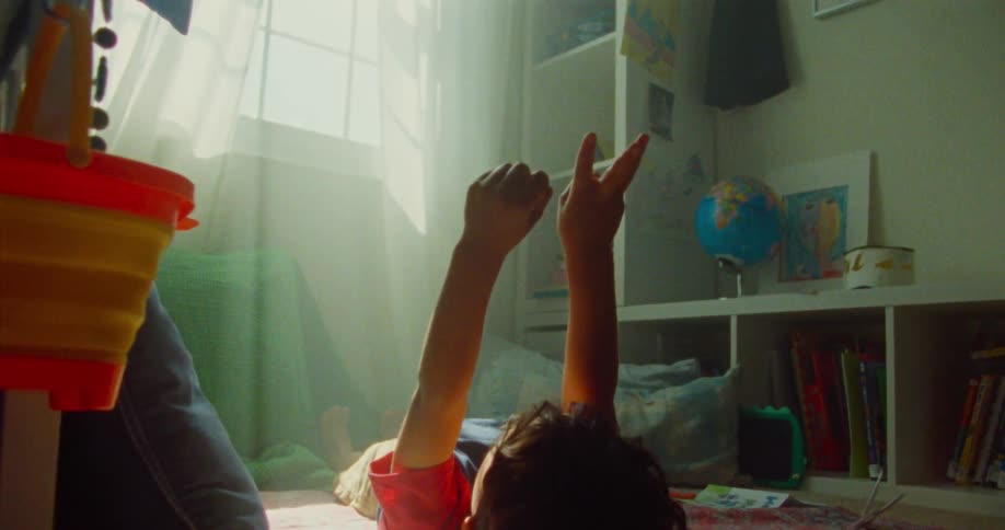 A child lies on a patterned rug in a sunlit room with arms raised and shelves filled with books and a globe visible in the background