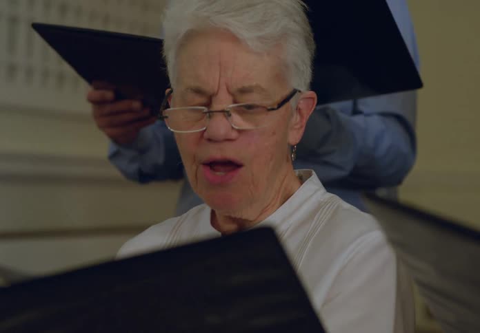 An elderly woman is singing from sheet music in a choir group