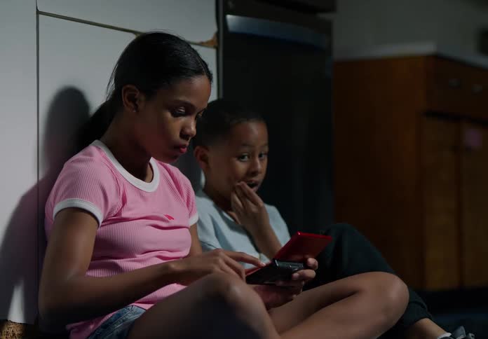 Two children sit on the floor against a wall eating snacks and looking at a handheld video game console