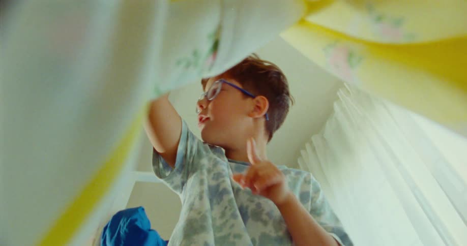A child wearing glasses and a patterned shirt lifts a blanket looks down and places a finger to their lips as if requesting silence