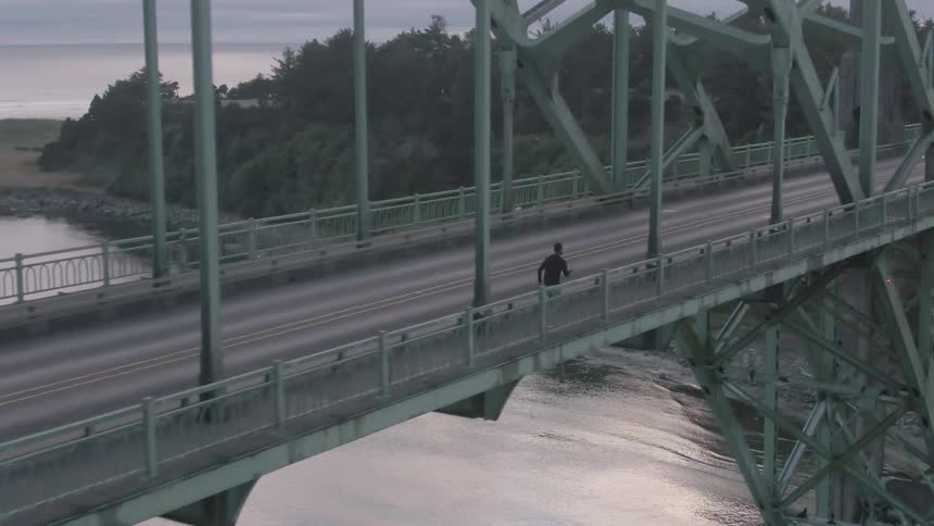 The man is jogging across the bridge as traffic goes by