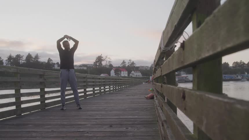 A man stands on a wooden bridge at midday stretching his shoulder and arm