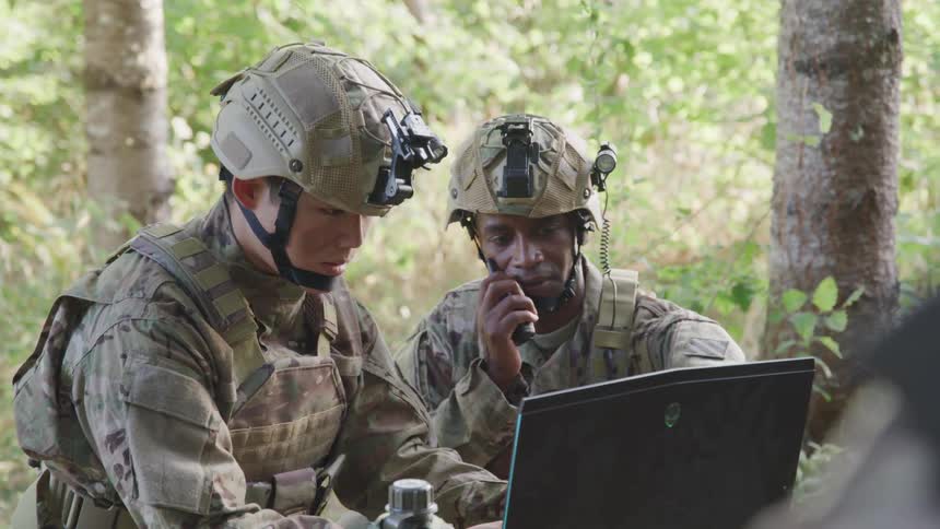 Two soldiers in camouflage uniforms and tactical gear are working on a laptop in a forested area