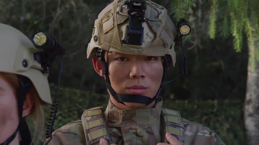 A man in a fully kitted military outfit wearing a helmet and holding the strap of a backpack on his shoulder stands next to another soldier outdoors