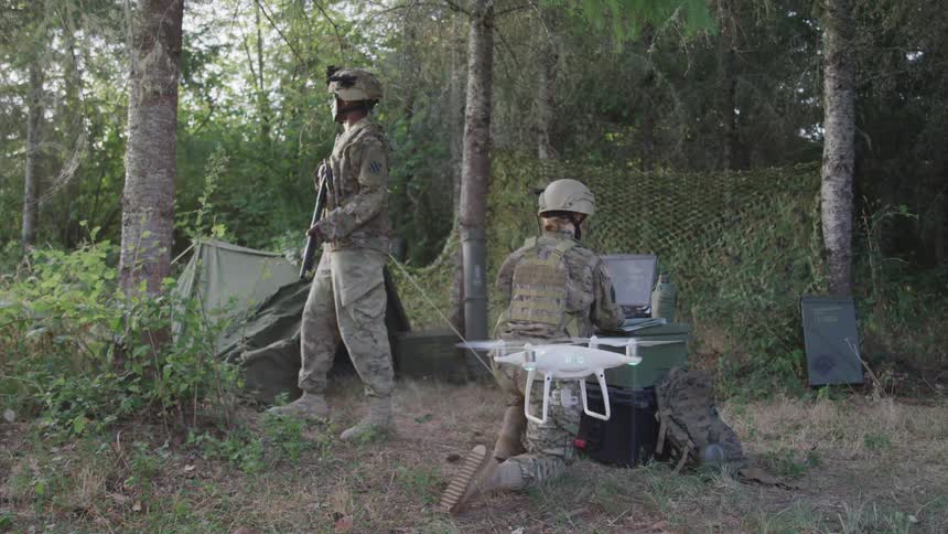 Two military officers are in the jungle at midday one operates a drone using a computer while the other stands guard