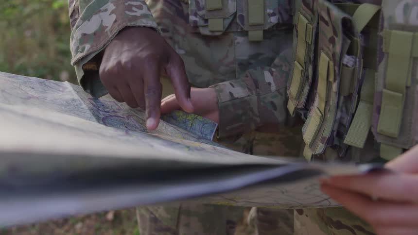 Two individuals in camouflage uniforms examine a map outdoors with one pointing to a location on it
