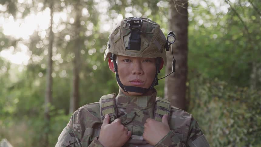 A young male soldier stands with his hands on his chest rig while wearing a helmet and looking forward in an outdoor setting with a forest in the background