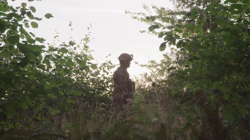 A military person wearing a uniform and helmet stands alone in a bush at midday
