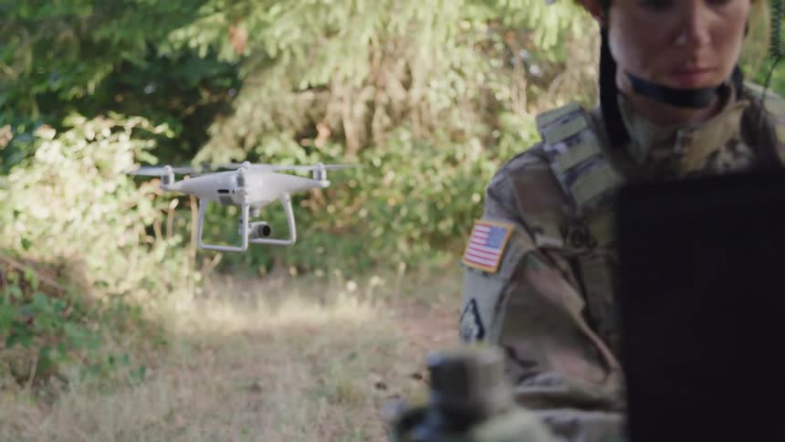 A soldier in camouflage uniform operates a drone in a forested area