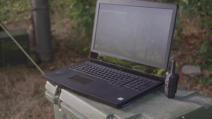 A laptop computer sits on a trashcan out on a lawn next to a walkietalkie at midday