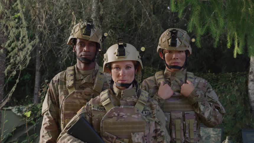 Three military personnel in uniform and with weapons in a camp in the forest