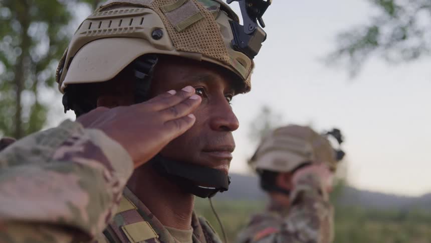 A black soldier standing outdoors next to another soldier salutes a superior at midday while positioned in a jungle setting