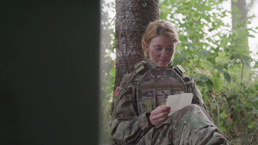 A blonde woman in soldier gear is sitting leaning against a tree reading a paper