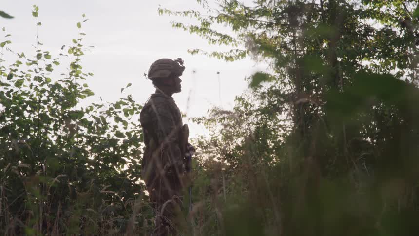A military officer stands armed in the jungle at midday wearing a helmet