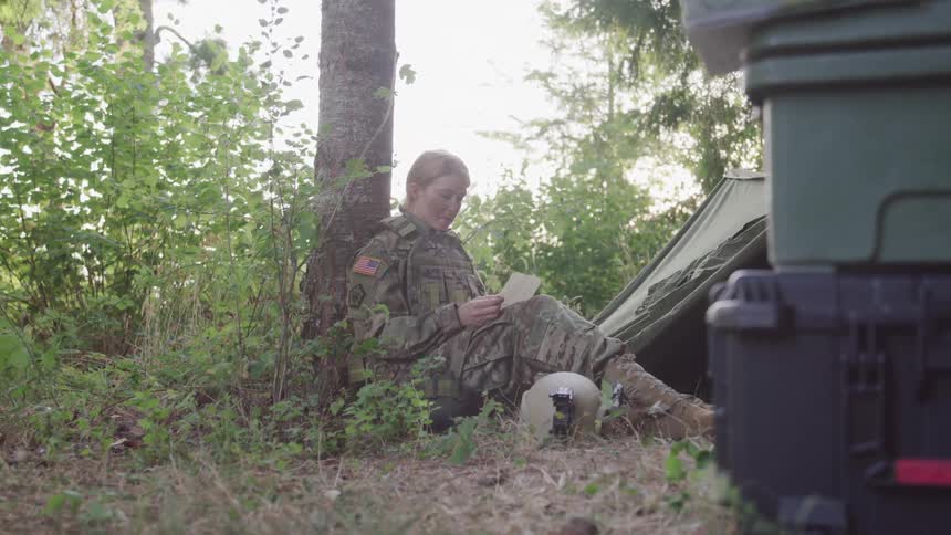 Military woman sitting in a tree in a makeshift camp reading a letter