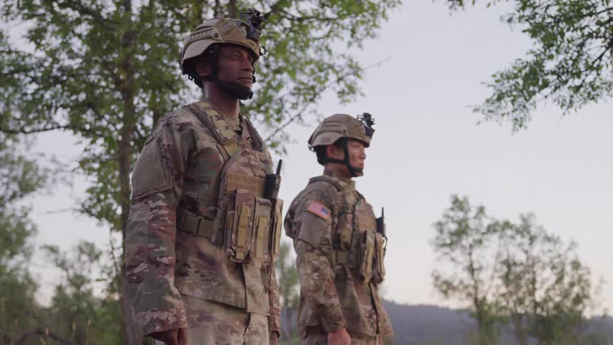 Two soldiers in camouflage uniforms and tactical gear stand outdoors with one wearing a helmetmounted camera and the other displaying a us flag patch on the arm