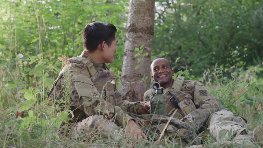 Two soldiers sit and lie on the grass in the jungle at midday laughing and talking as a young asian man sips water from a canteen